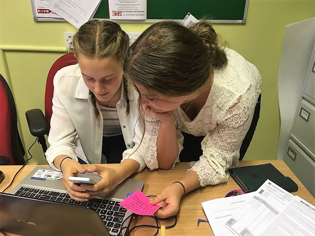 Two young women sit in front of a laptop and one types into a mobile phone on Digital Teen Takeover.