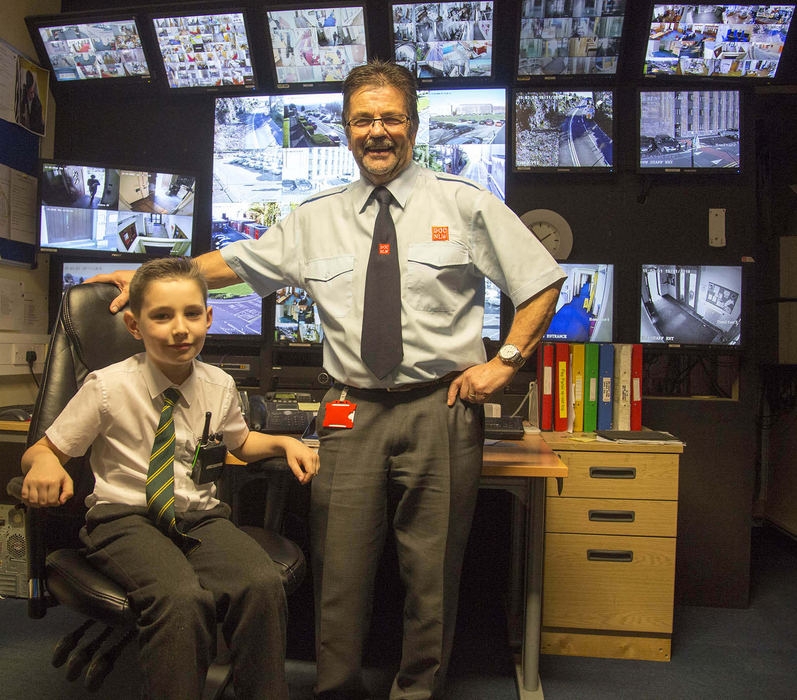 A young boy sits in a chair next to a security guard in front of CCTV screens at the National Library of Wales on Takeover Day.