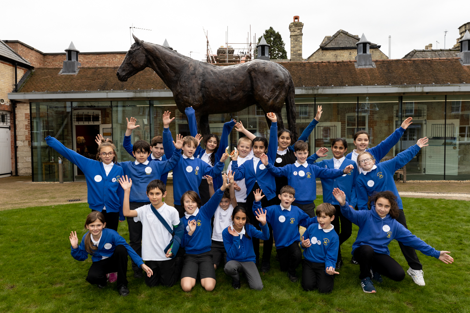 A group of uniformed primary school children grinning and holding their hands up in the air in front o a horse statue at the National Horseracing Museum.