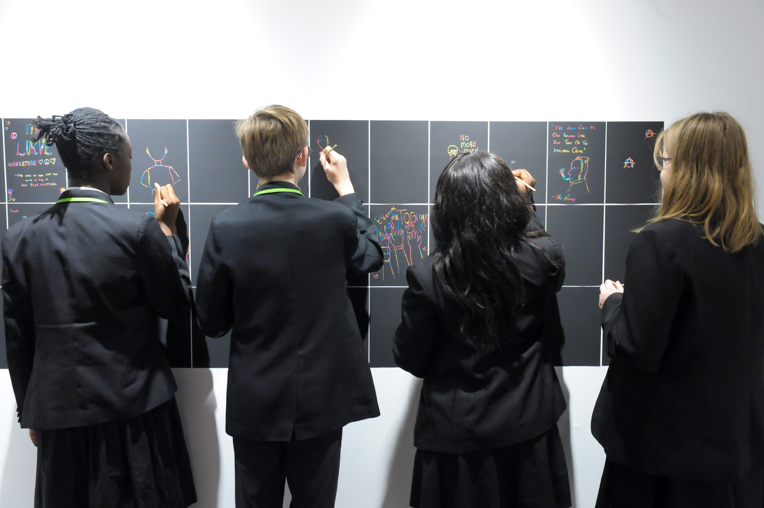 Four young people face the wall and draw on a chalkboard sign with their messages at the National justice museum.