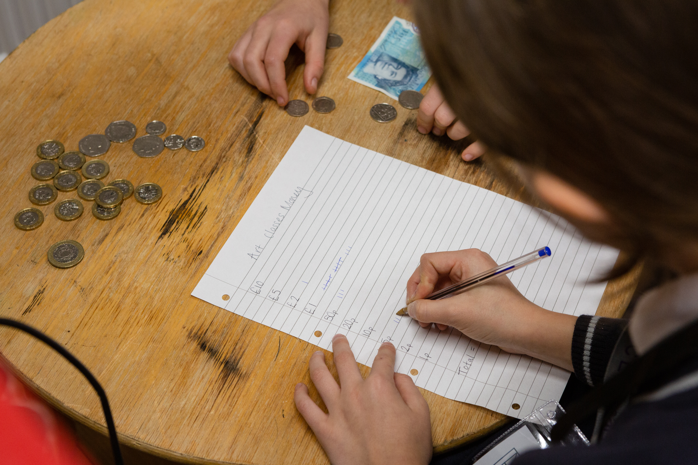 A school child leans on a desk covered with coins and writes on a piece of paper with the heading 'Art Class money' during the Lauderdale House Takeover Day.