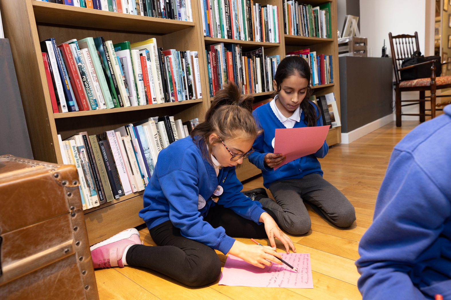 Two primary pupils sit on the floor and draw on pink pieces of card in the National Horseracing Museum shop.