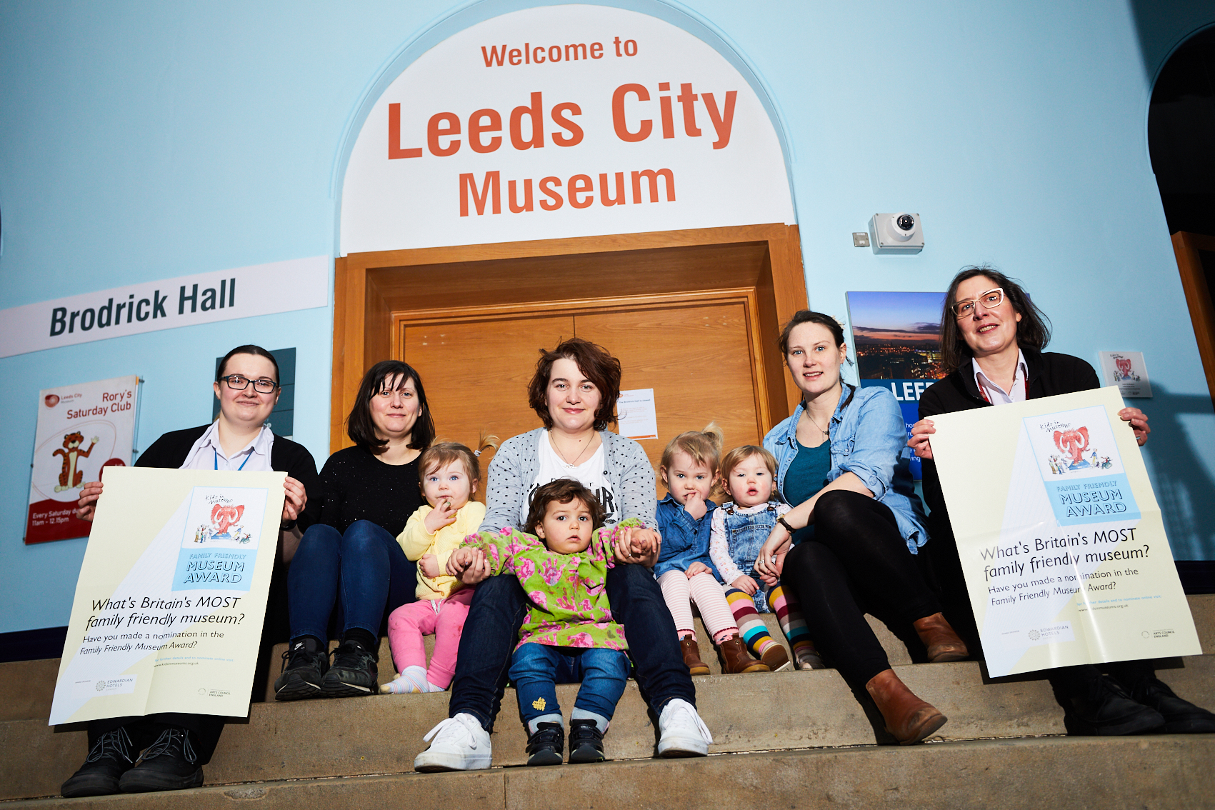 Museum staff, toddlers and parents sit on the steps inside Leeds City Museum reception holding Family Friendly Museum Award posters.