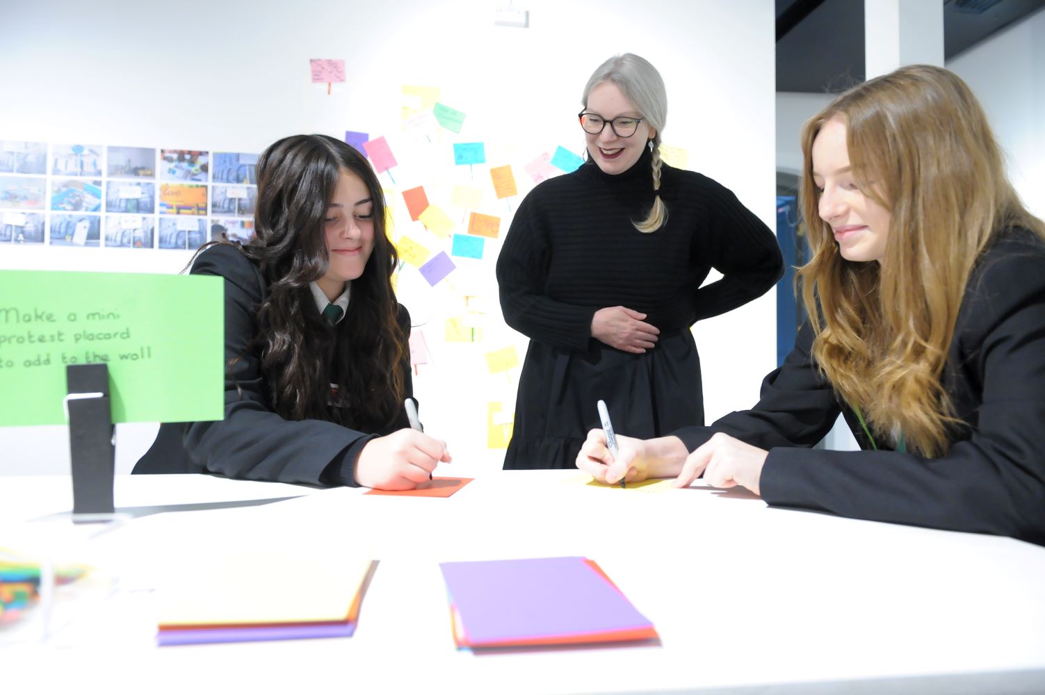 Two teenagers sit writing on post-it notes at a desk at the National Justice Museum in Nottingham. A member of museum staff stands in the background smiling.