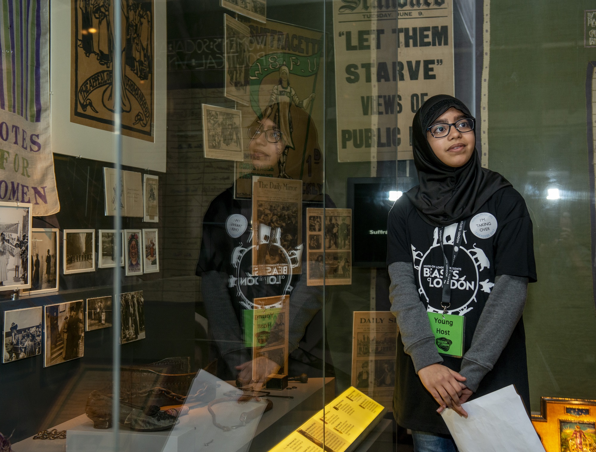 A young woman wearing a badge saying 'Museum Host' and 'I'm taking over' stands in front of a display case at the Museum of London.