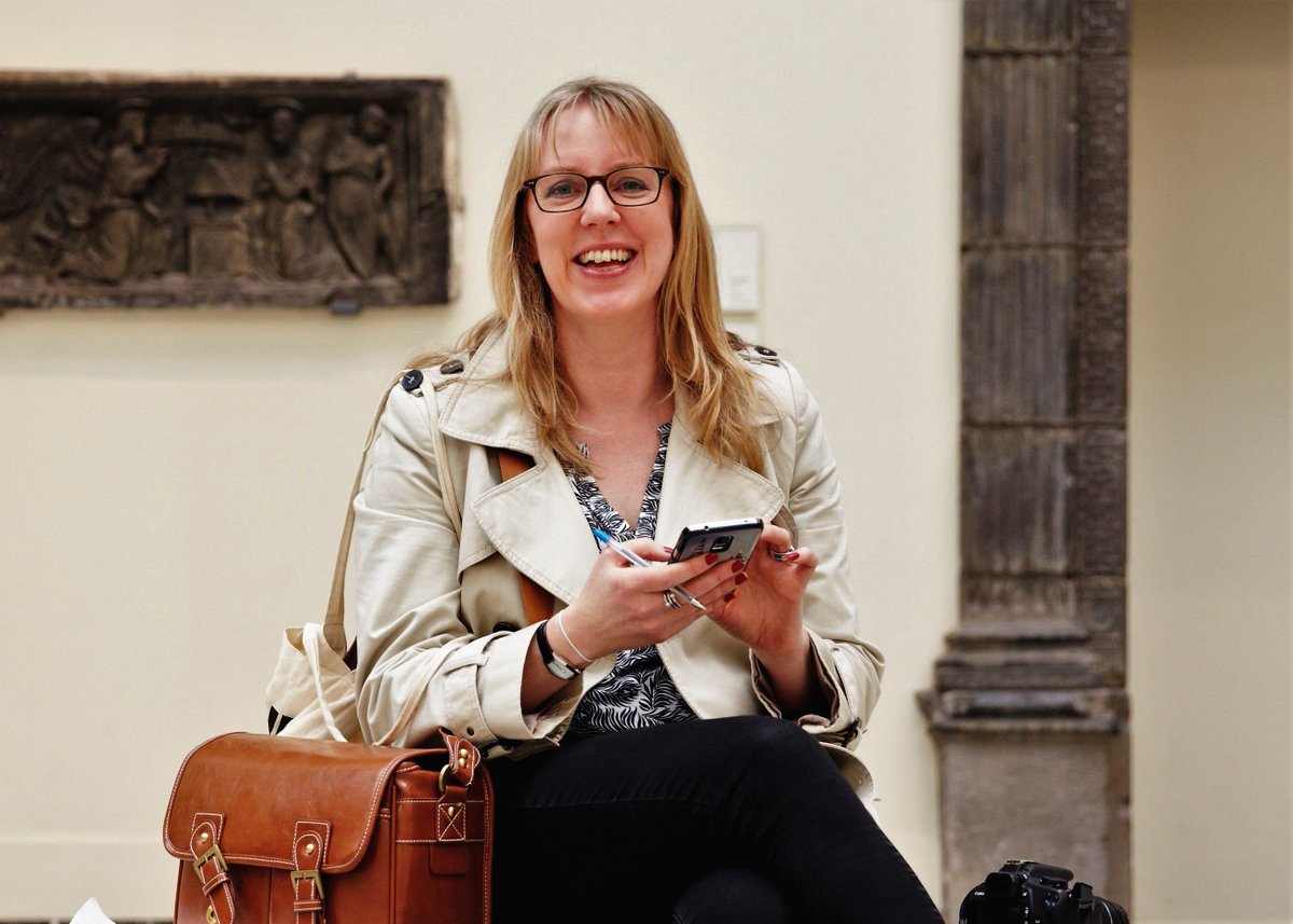 Claire Madge sits in the foyer of a museum smiling and holding her phone and a pen.
