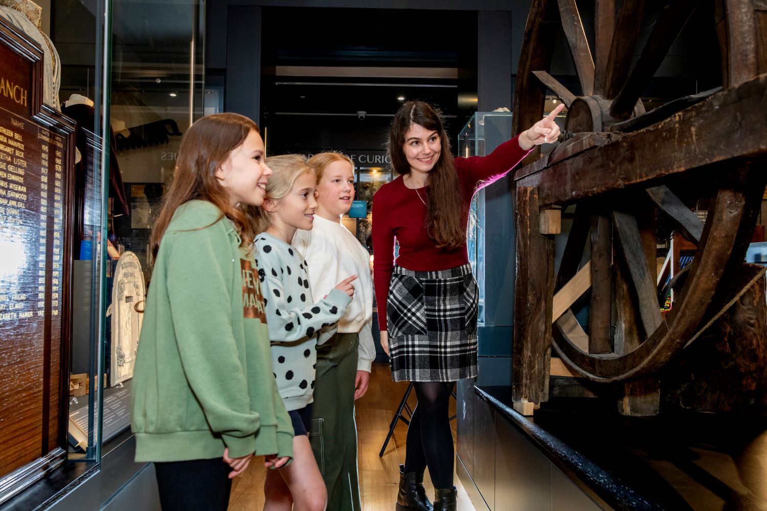 A member of museum staff points out a museum object to three young visitors at Craven Museum. They are stood in front a giant wooden wheel.