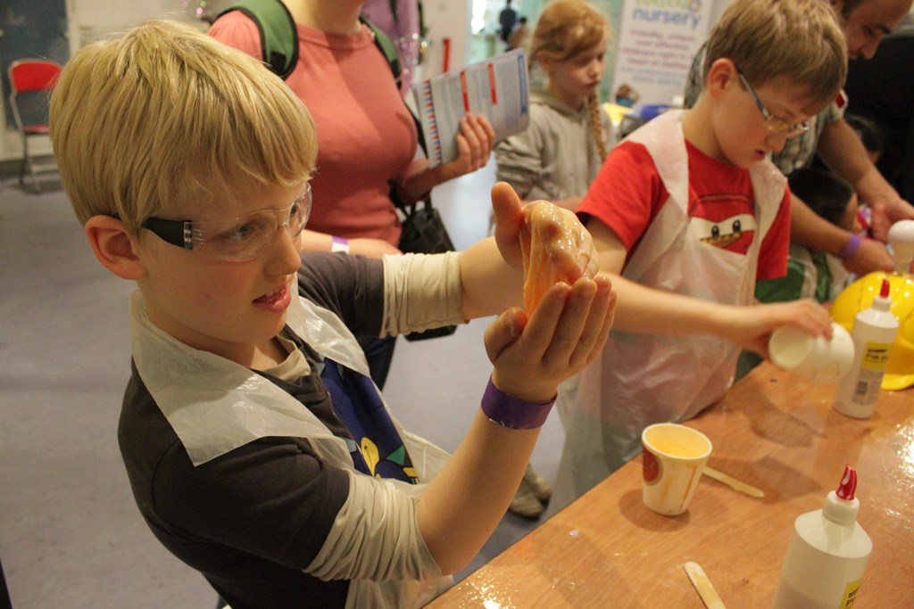 A young boy wearing goggles makes slime in a workshop with other children.