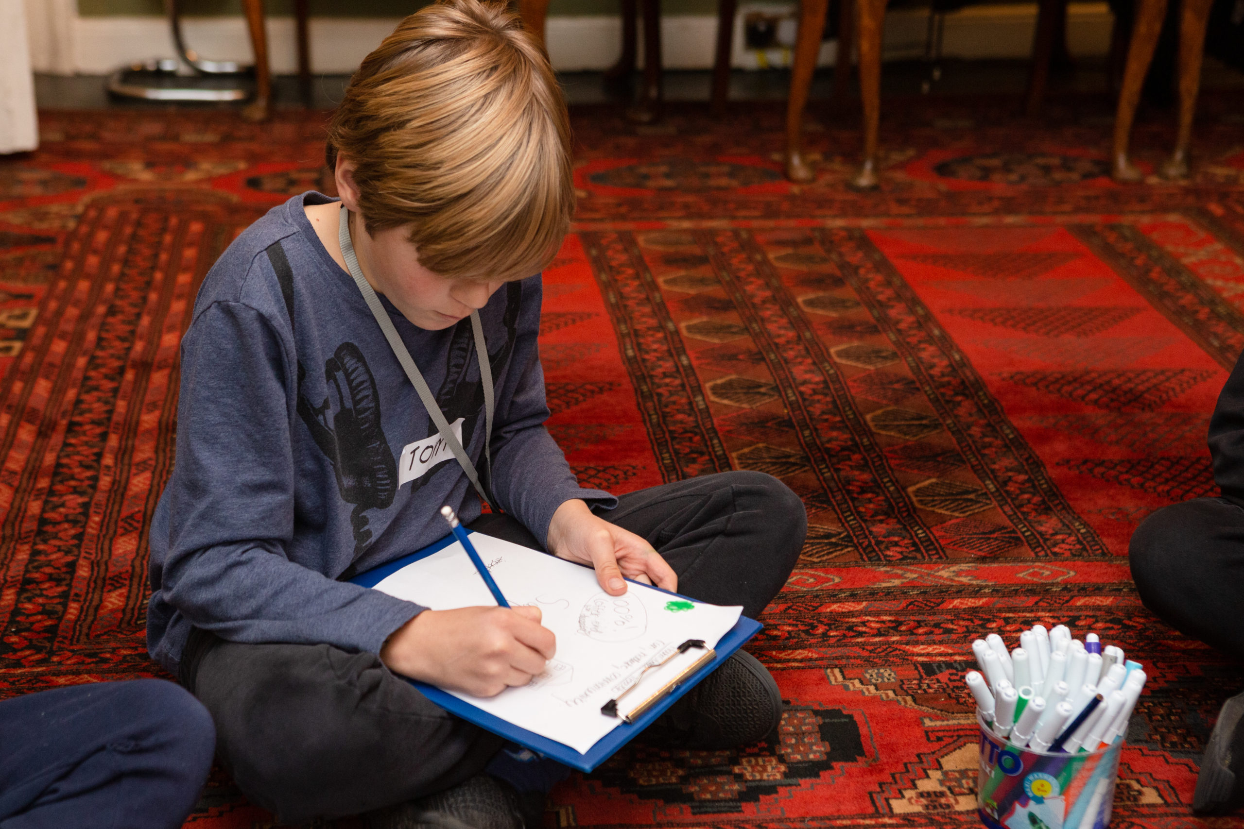 A young boy draws on a clipboard on a carpeted floor.