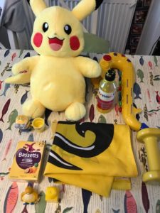 A table covered in yellow objects, including a yellow Pikachu teddy, paint, blow up saxophone, vitamins and various small toys.