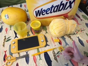 A selection of yellow objects laying on a table, including a gameboy, Weetabix, wool, cups and a melon.