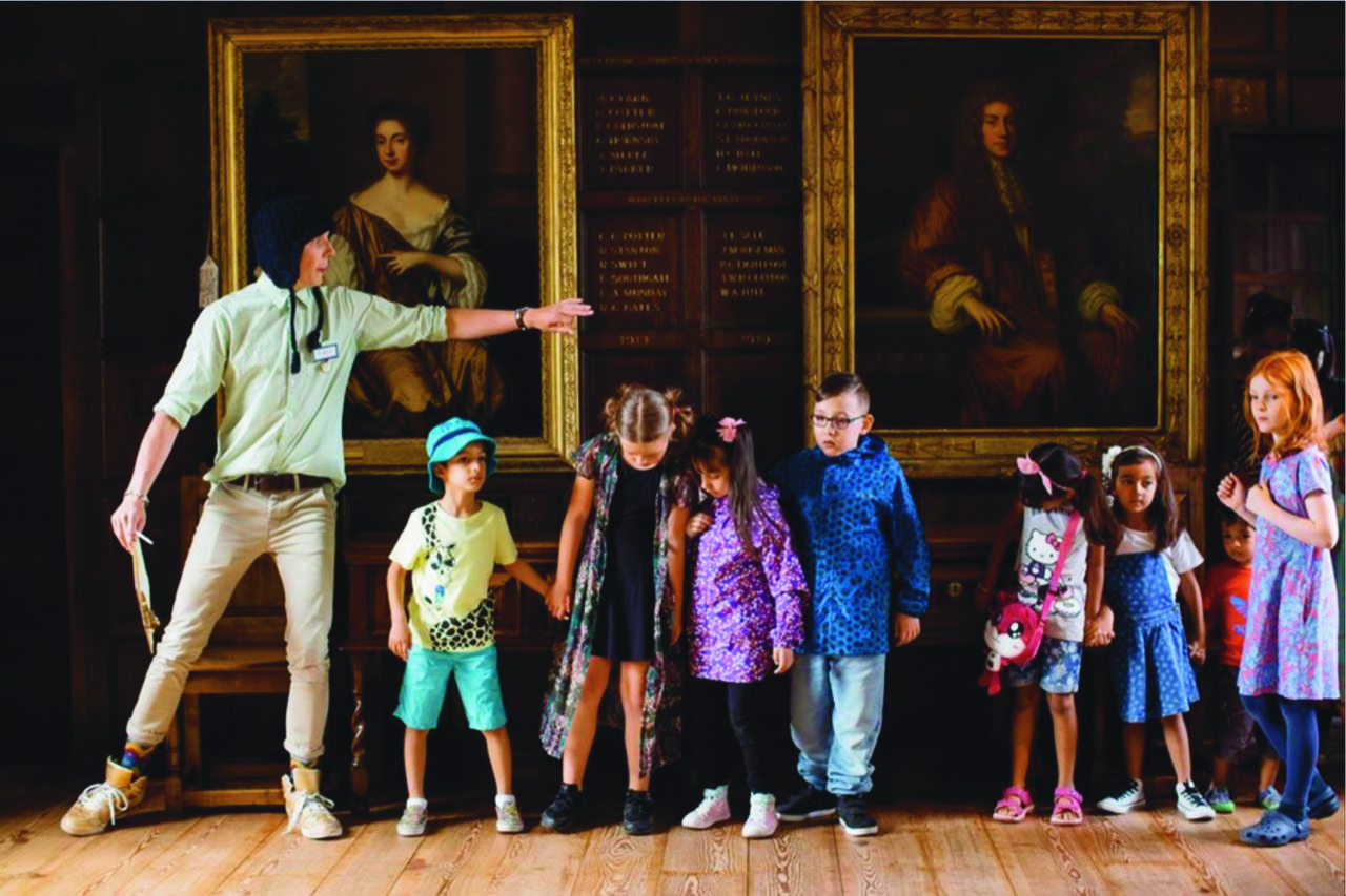 An adult leads a group of children stood across a gallery in front of a wall of portraits.