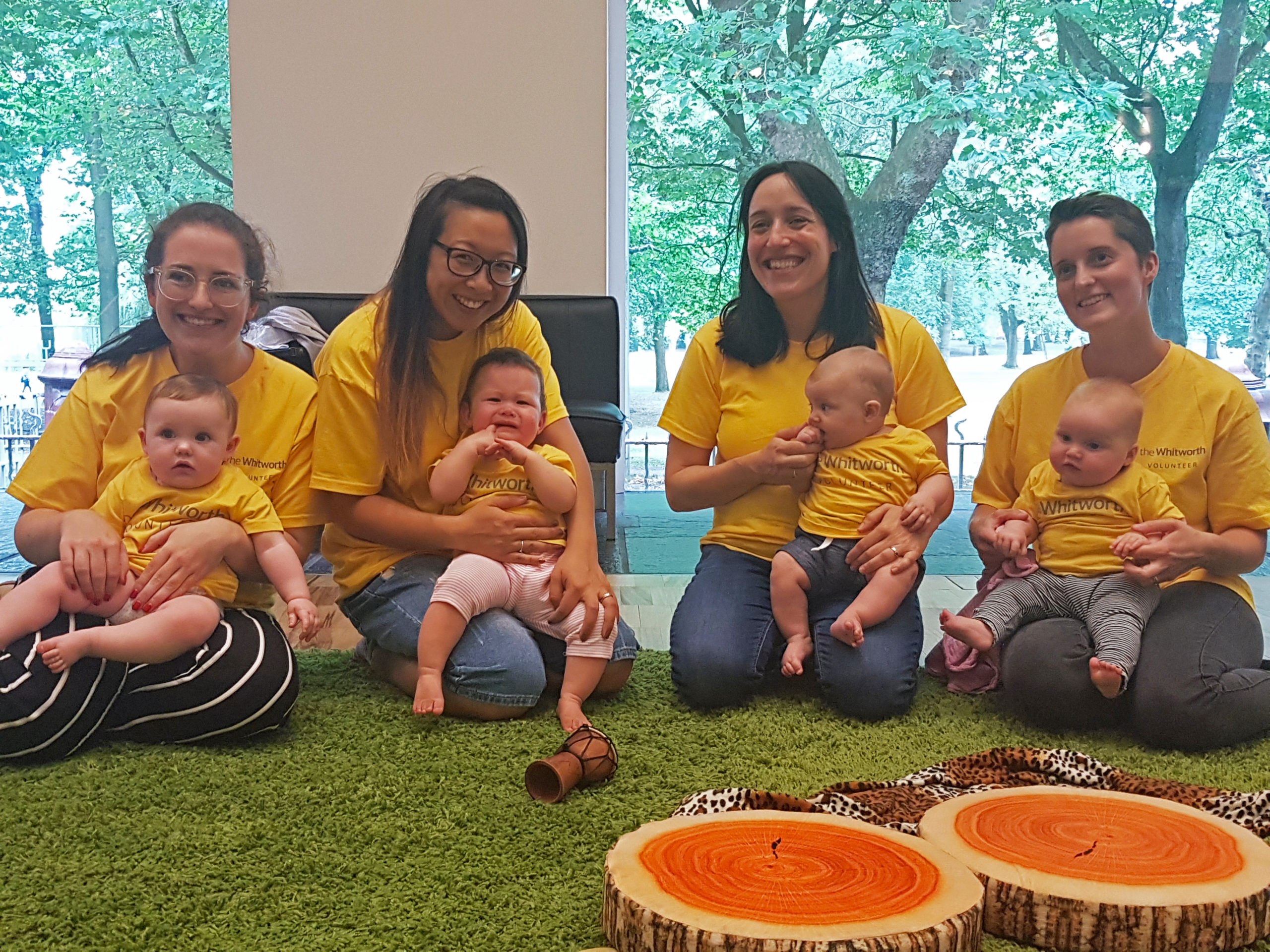 Four women kneel holding a baby each on their laps all wearing yellow volunteer t-shirts.
