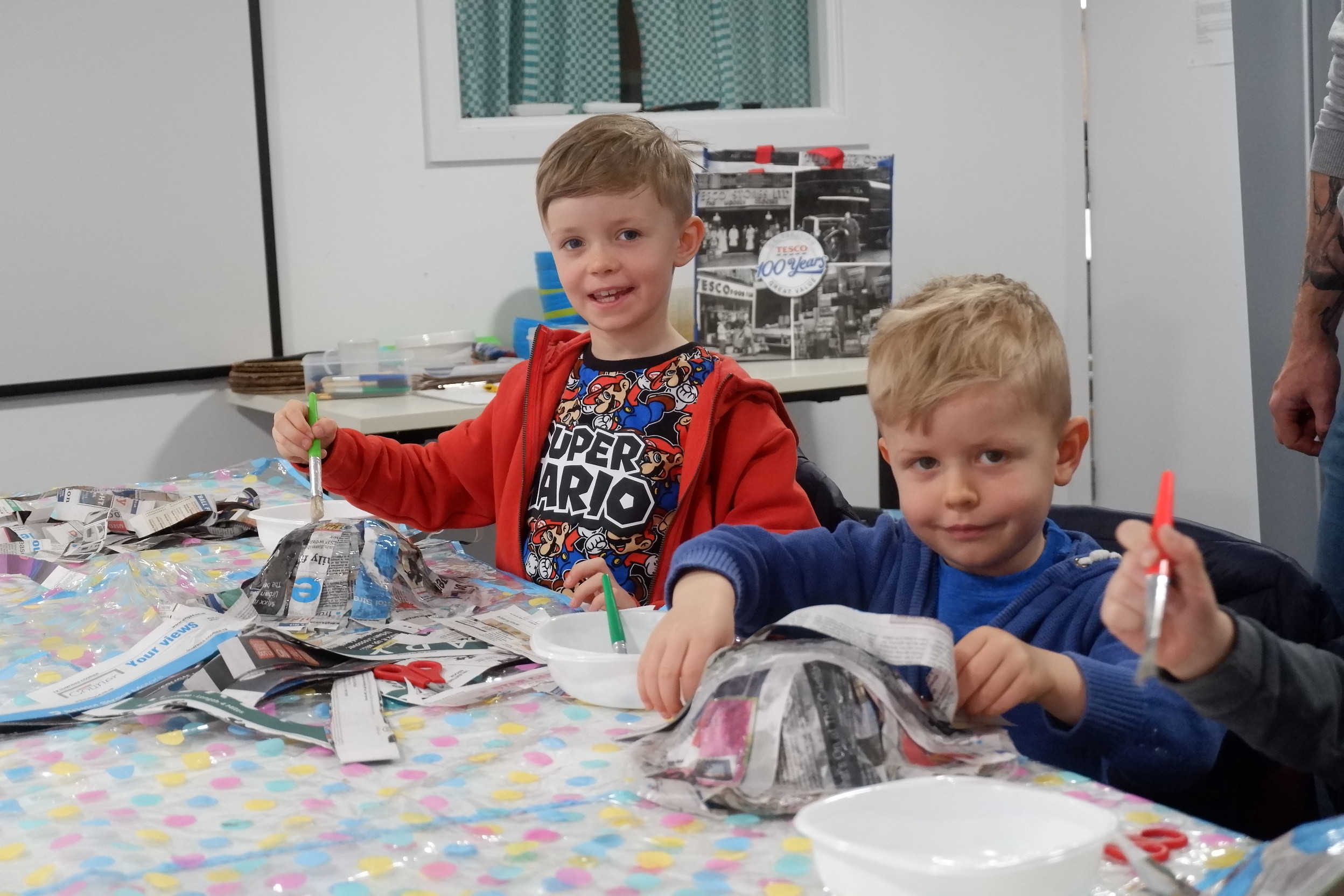 Two young boys smile at the camera as they make paper maché helmets sat at a table.