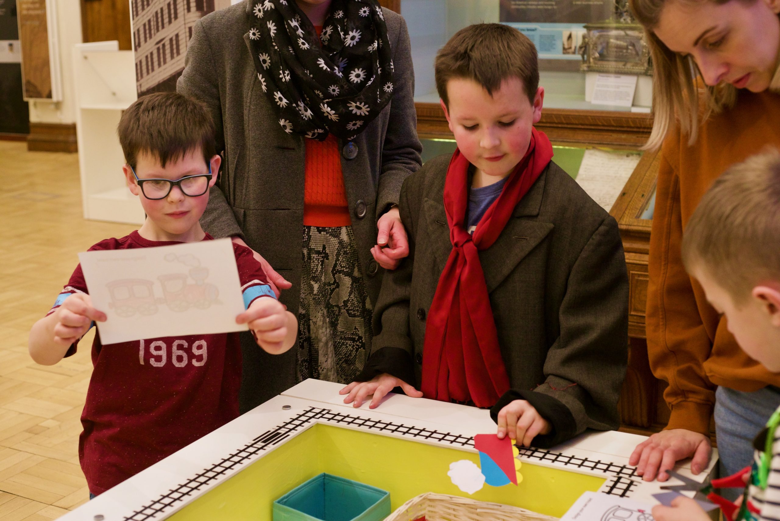 Three young boys colour in pictures of trains at a yellow craft station at the Andrew Carnegie Birthplace Museum with their mothers stood behind them.