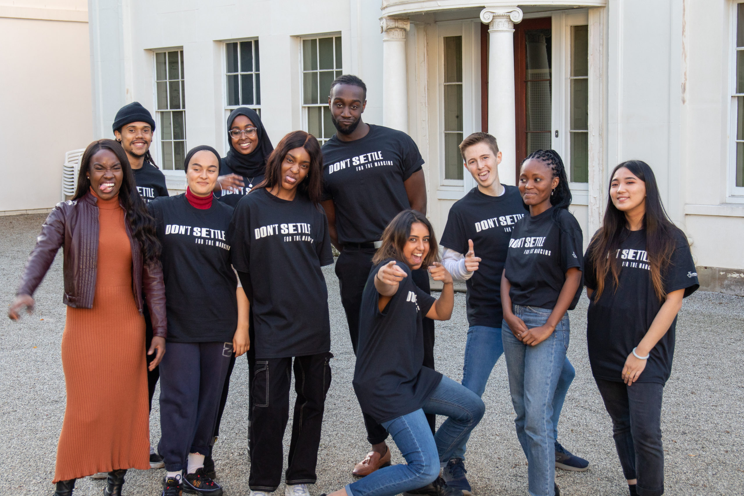 A group of young people wearing black t shirts reading Don't Settle in white text smiling and standing in front of a historic white building.