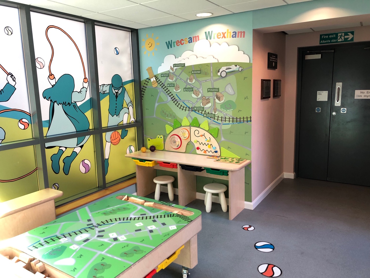 A play room in Wrexham Museum with map mural, stained glass window of children playing, play table and stools.