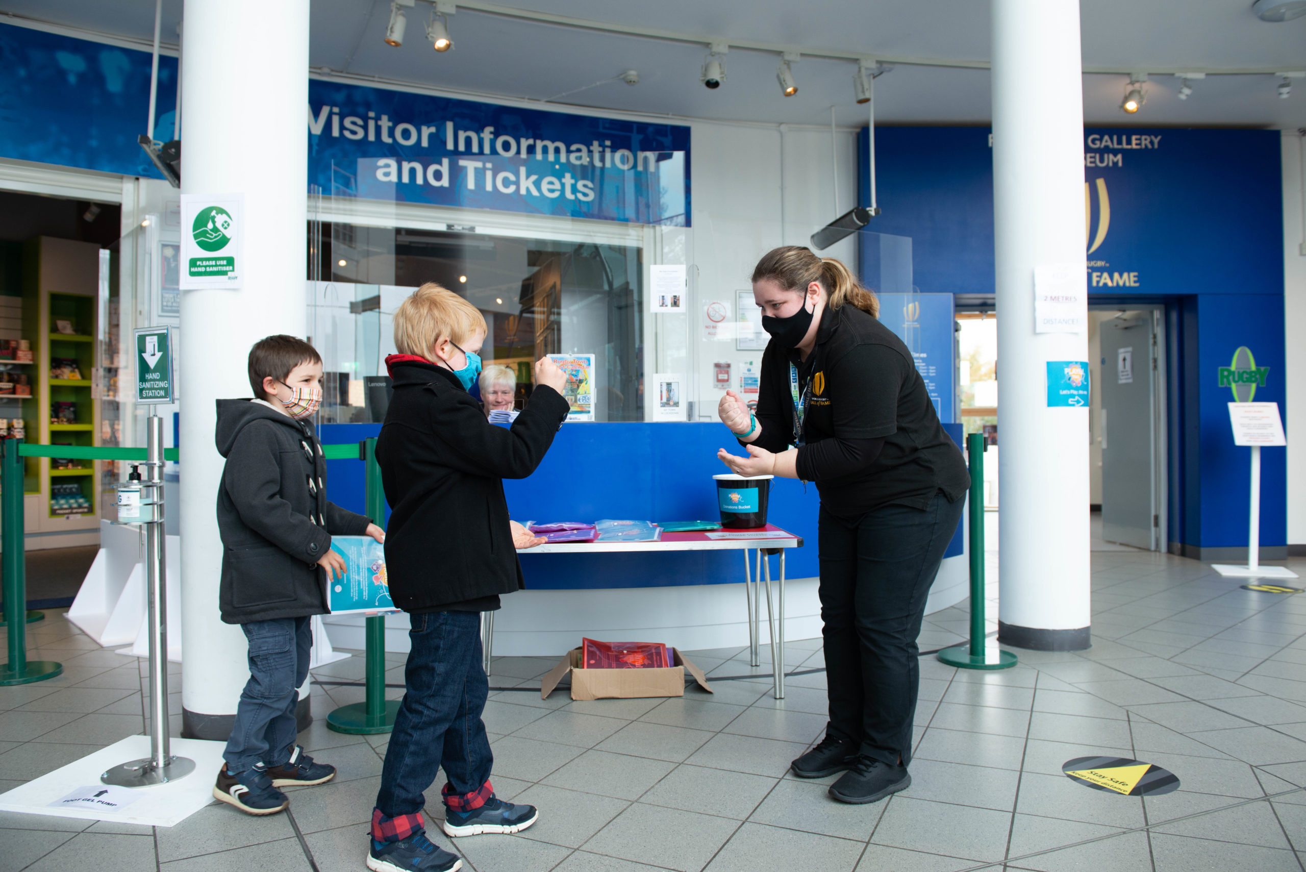 A museum staff member plays rock paper scissors with two young boys in the entrance hall of Rugby museum.