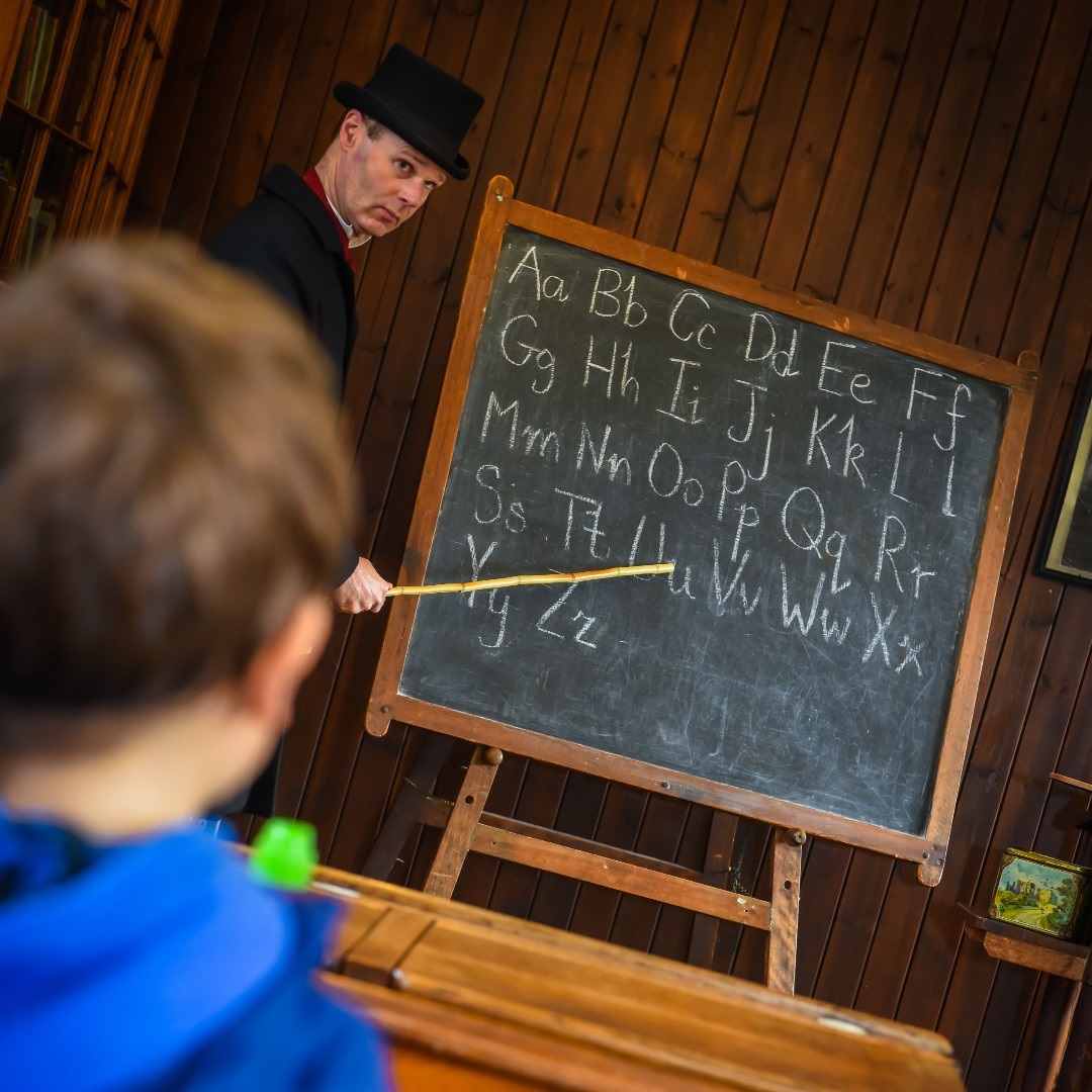 A stern looking Victorian schoolmaster wearing a top hat points with a stick at a blackboard with the alphabet written on it. A young boy in a blue hoodies sits facing towards the board.