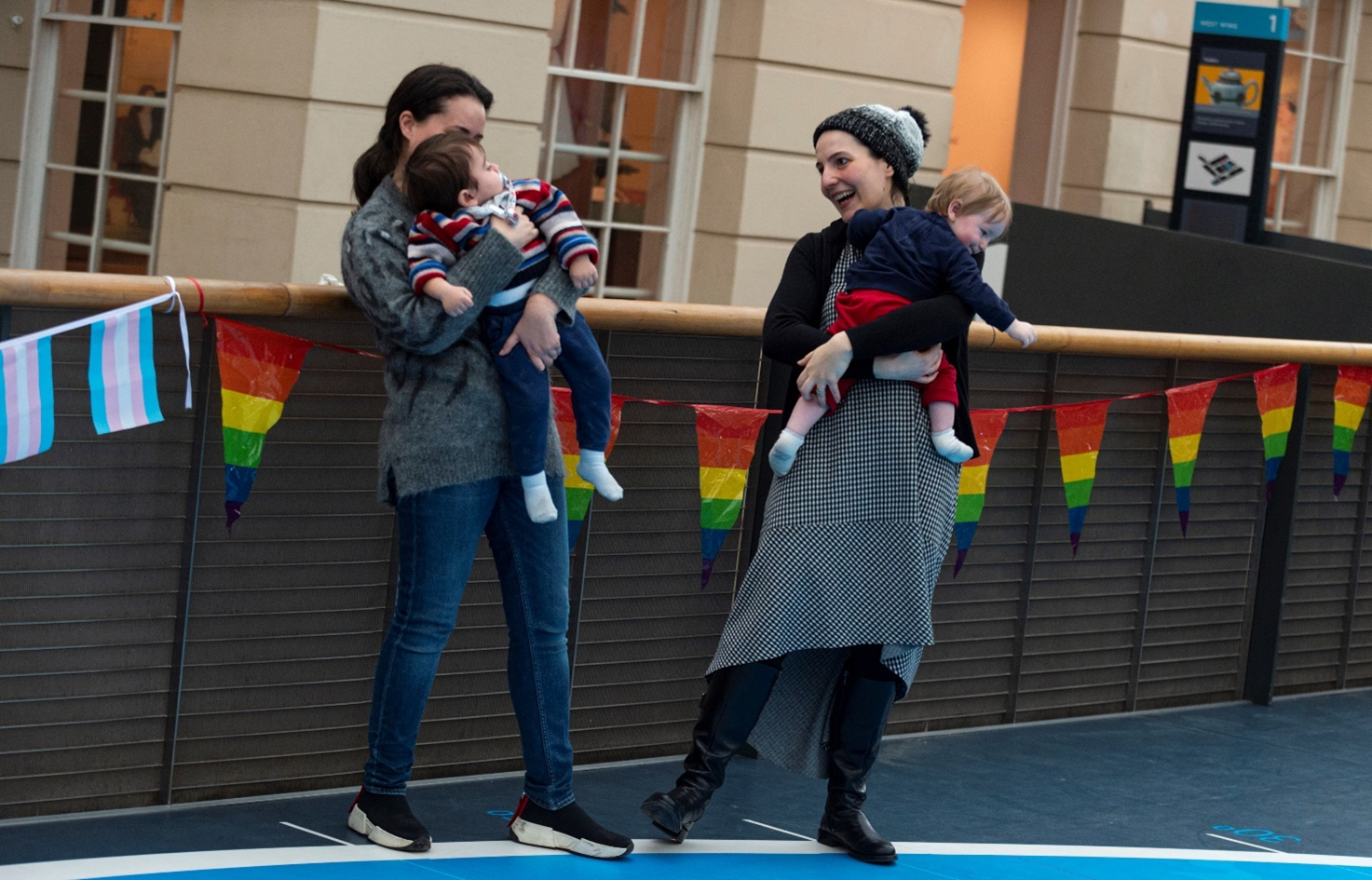 Two women holding toddlers in their arms in a museum gallery with rainbow flags hung up behind them.