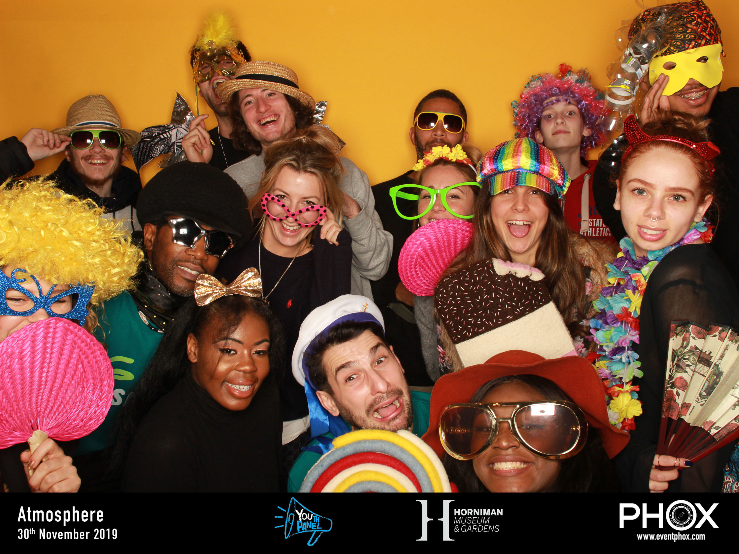 Members of the Kids in Museums Youth Panle pose in a photo booth with a yellow background with different props like big sunglasses, wigs and hats.