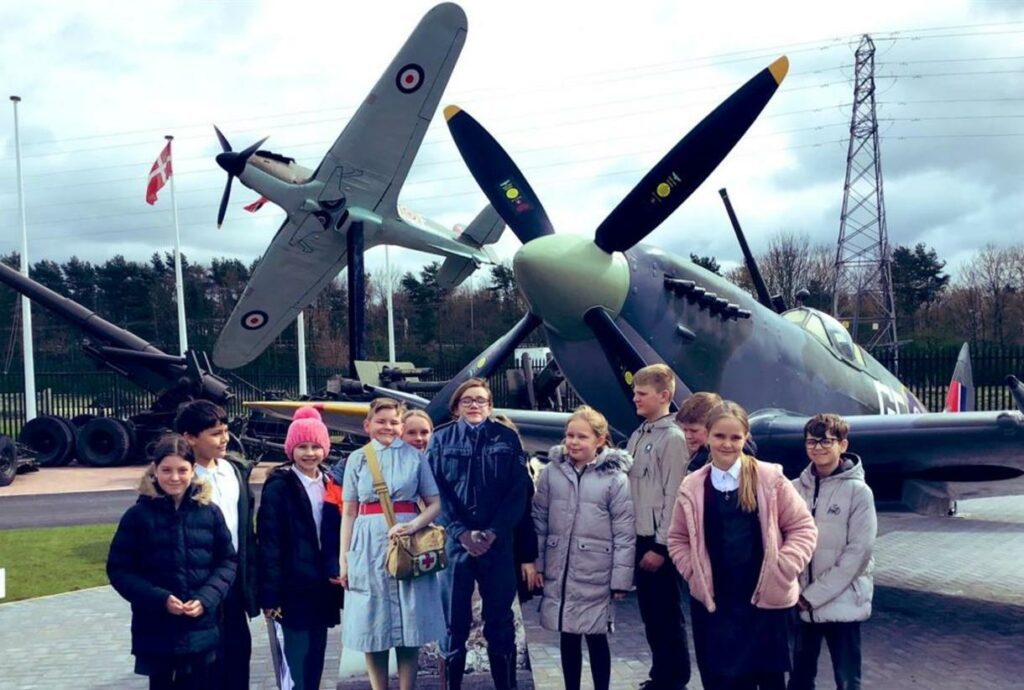 A group of school children stand in front of two World War Two planes. In the middle of the group, two children stand behind headless cutouts of a nurse and uniformed officer from the 1940s.