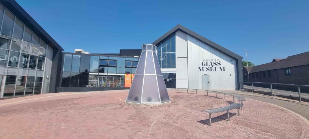 An exterior shot of Stonebridge Glass Museum on a bright sunny day. The building has many large, clear windows. In front of the museum is an upside down funnel shaped glass sculpture surrounded by benches 