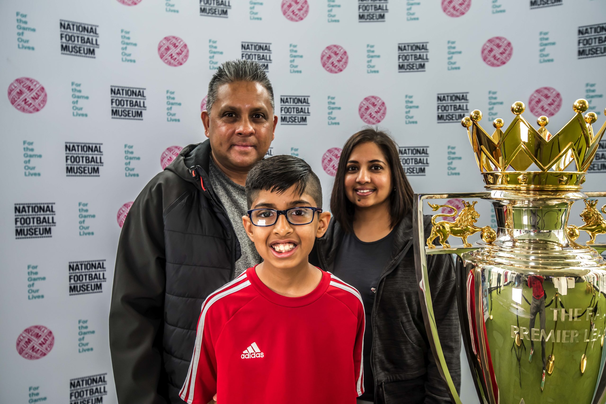 A boy and his parents stand in front of a premier league trophy at the national football museum.