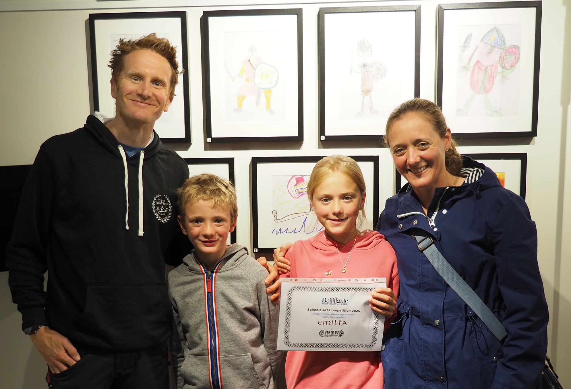 A family of two adults and two children, one holding a certificate, in front of a wall of framed children's pictures.