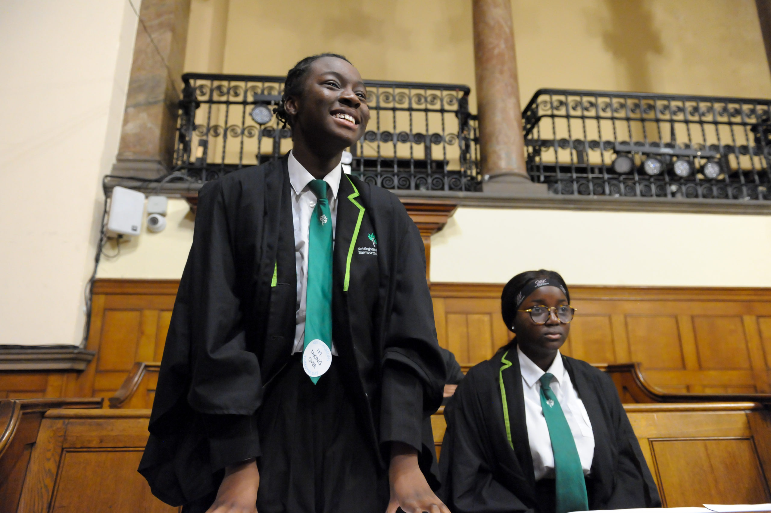 Two young women in the courtroom, the one nearest standing, smiling and wearing a takeover day sticker.