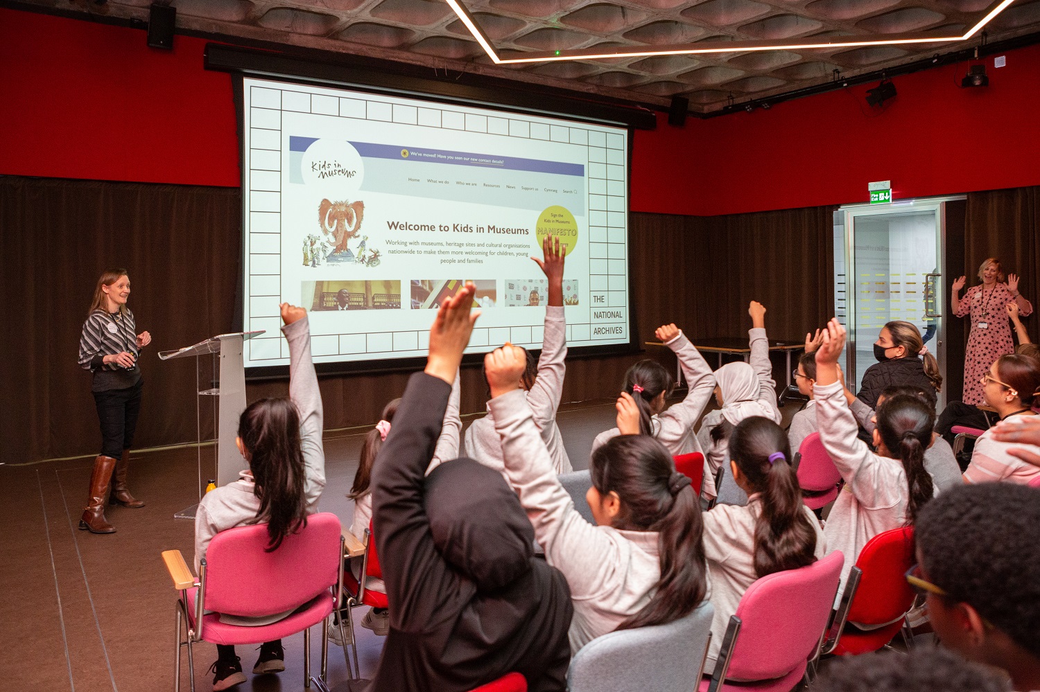 A woman stands next to a projector screen showing the Kids in Museums website in front a group of children all with their hands in the air at the national archives takeover day.