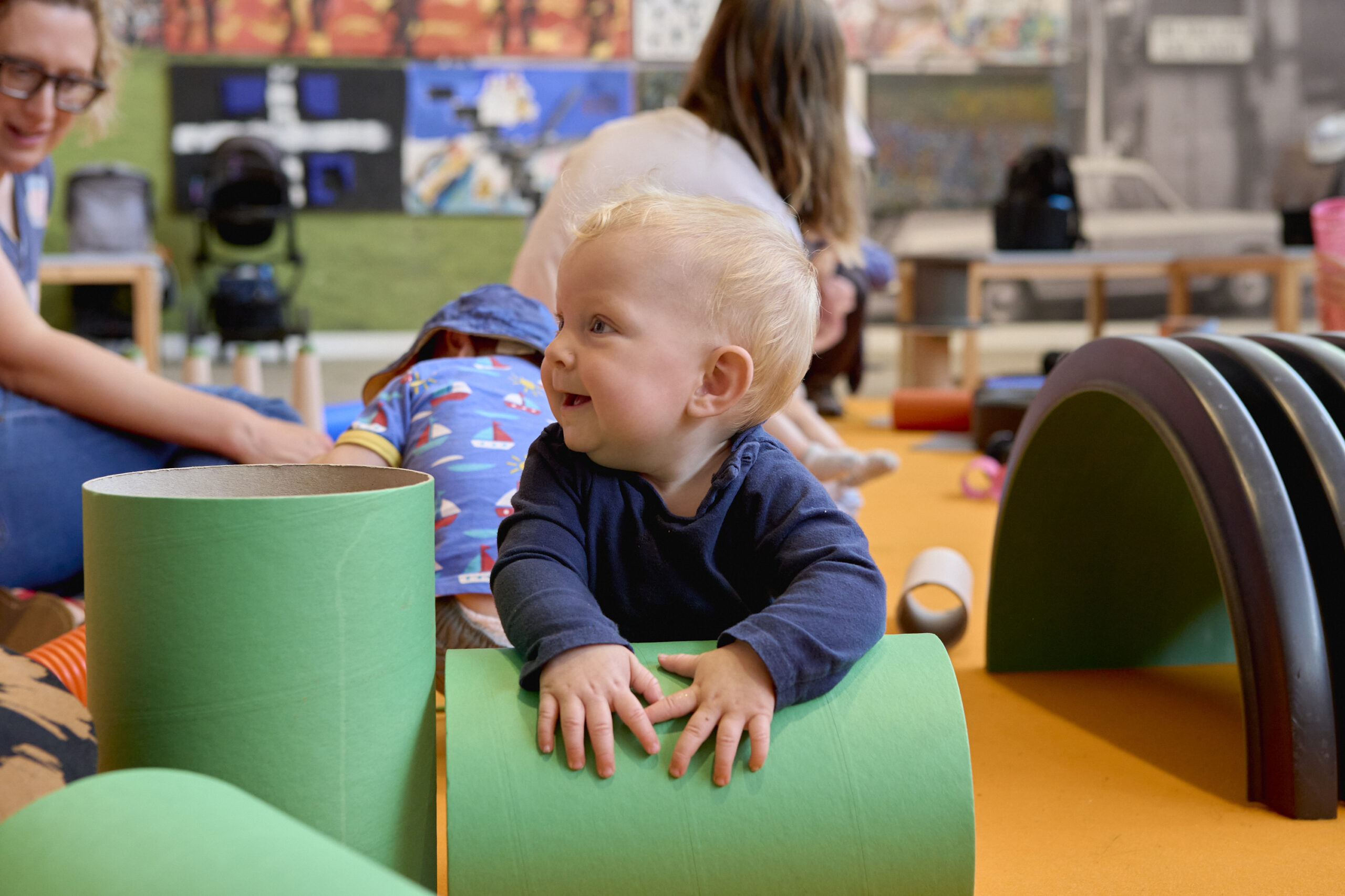 A baby in a blue long sleeved outfit leans on a large green tube during a play session at The Whitworth. Families can be seen playing with other play equipment in the background.