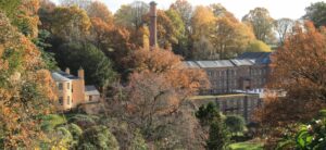 An industrial mill sits in the middle of autumnal trees. The building is long and made of brick, with rows of windows and a long chimney. There is a lighter coloured house to the left of the mill.