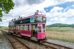 A pink double decker tram sits on a tramline against a background of rolling hills and fields. There are people sitting on the top deck and you can see the drive at the front of the tram.