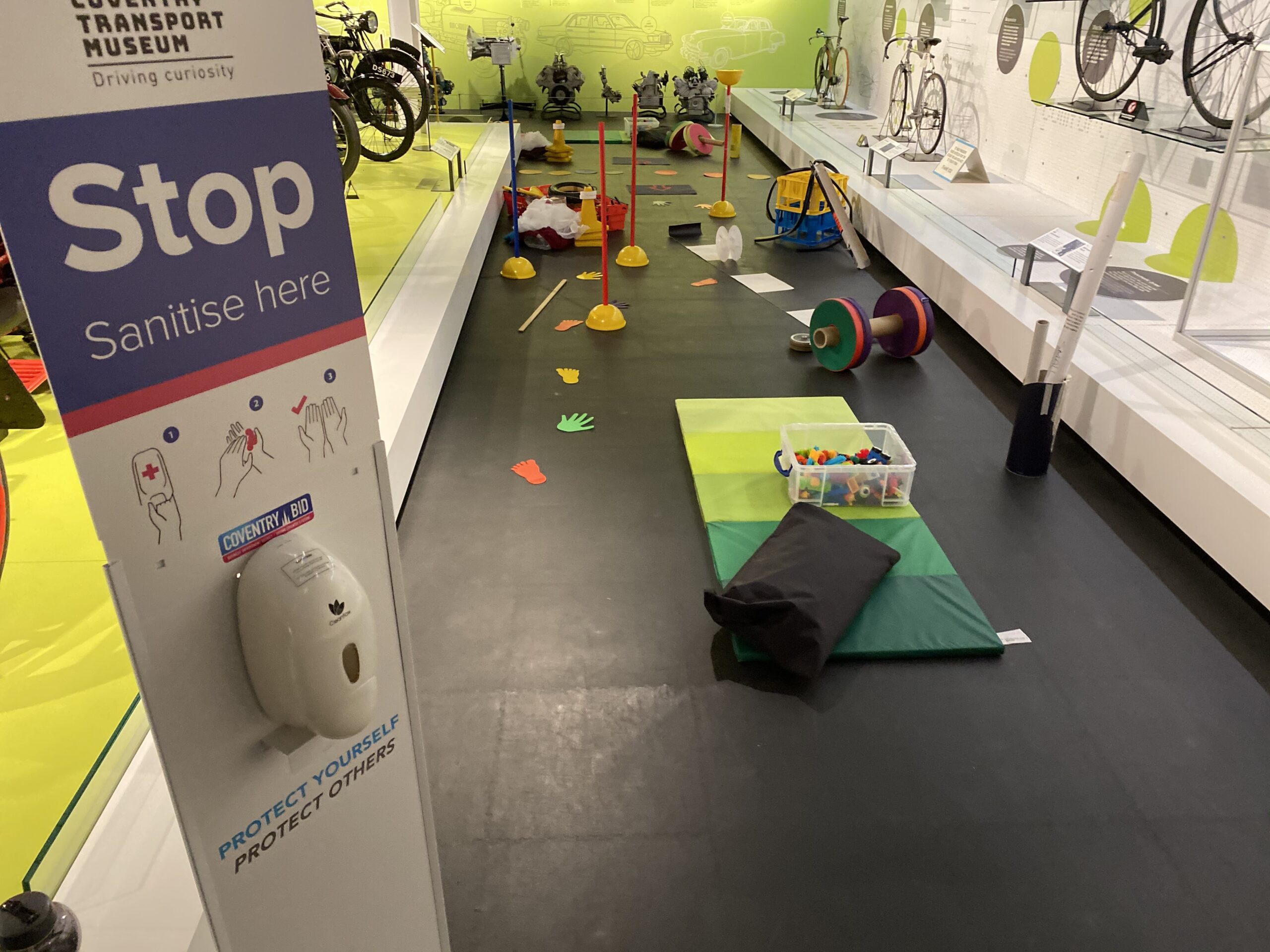 A playmat and various toys and loose parts are laid out on the floor in a gallery display of bicycles.