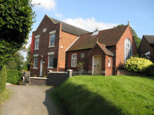A red brick chapel building with a grassy bank and path leading up towards the gate.