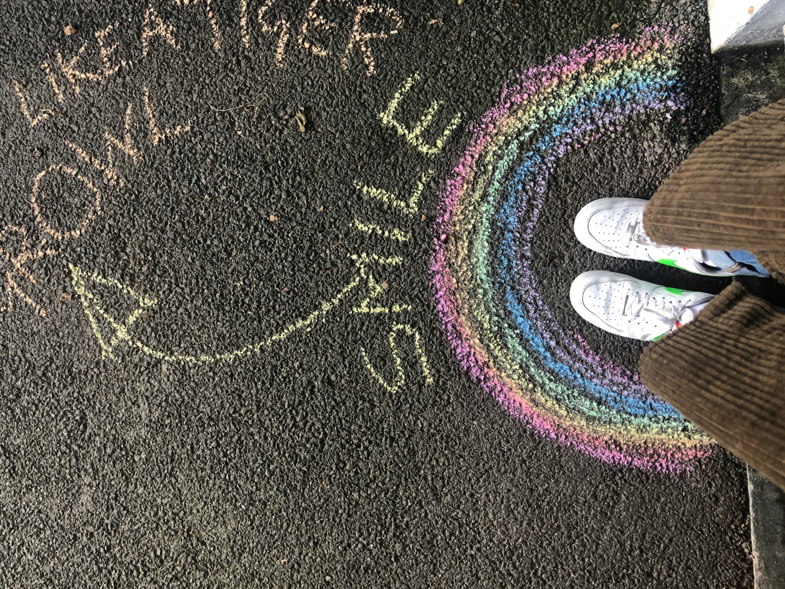 Close up of a hopscotch drawn in chalk on tarmac. There is a rainbow drawn on the right with somebody standing inside it.