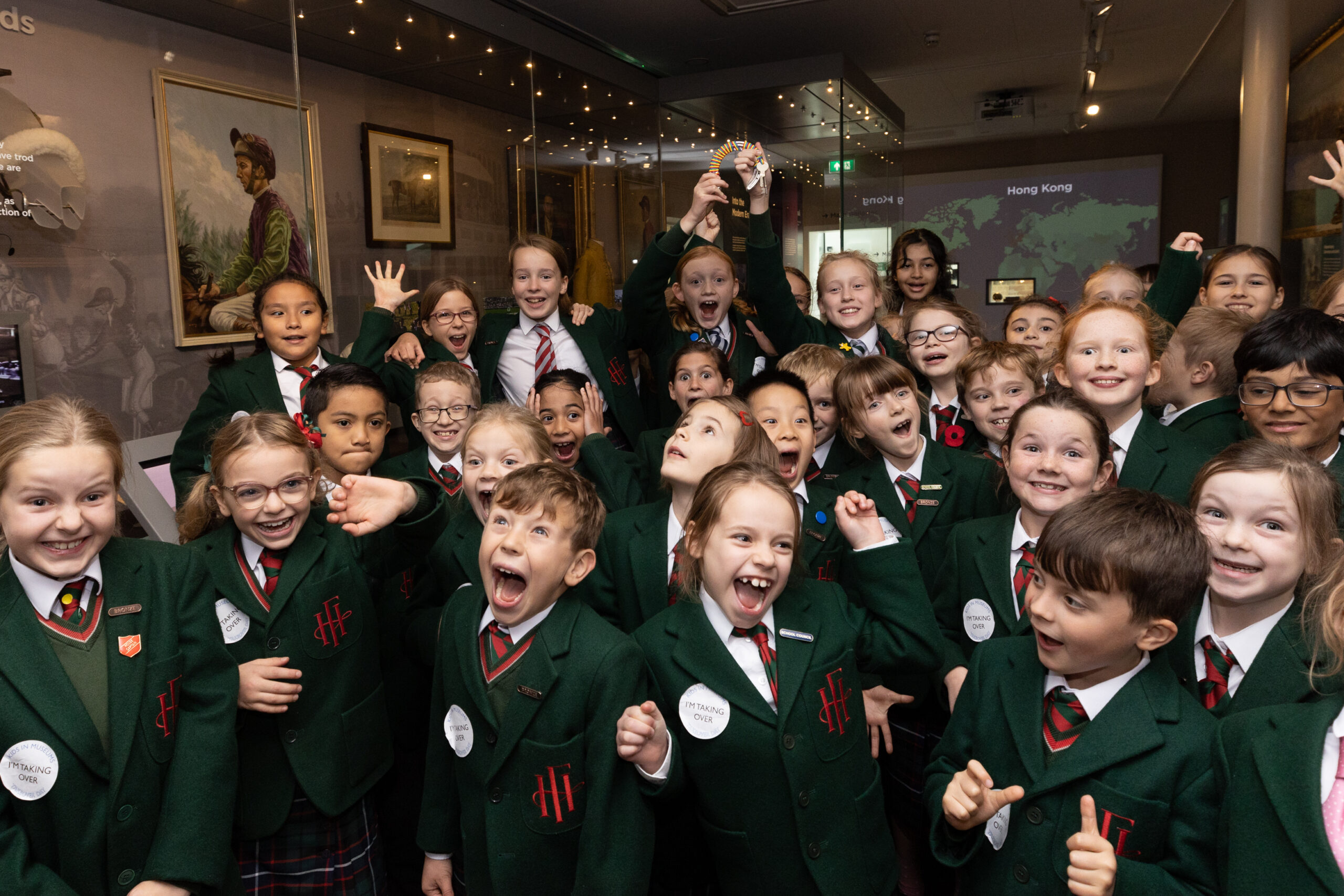 A group of excited grinning children inside a gallery at the National Horseracing Museum with glass museum cases behind them. Two children at the back of the image hold up a horseshoe shaped set of keys in the air. They are wearing stickers reading 'I'm taking over'.