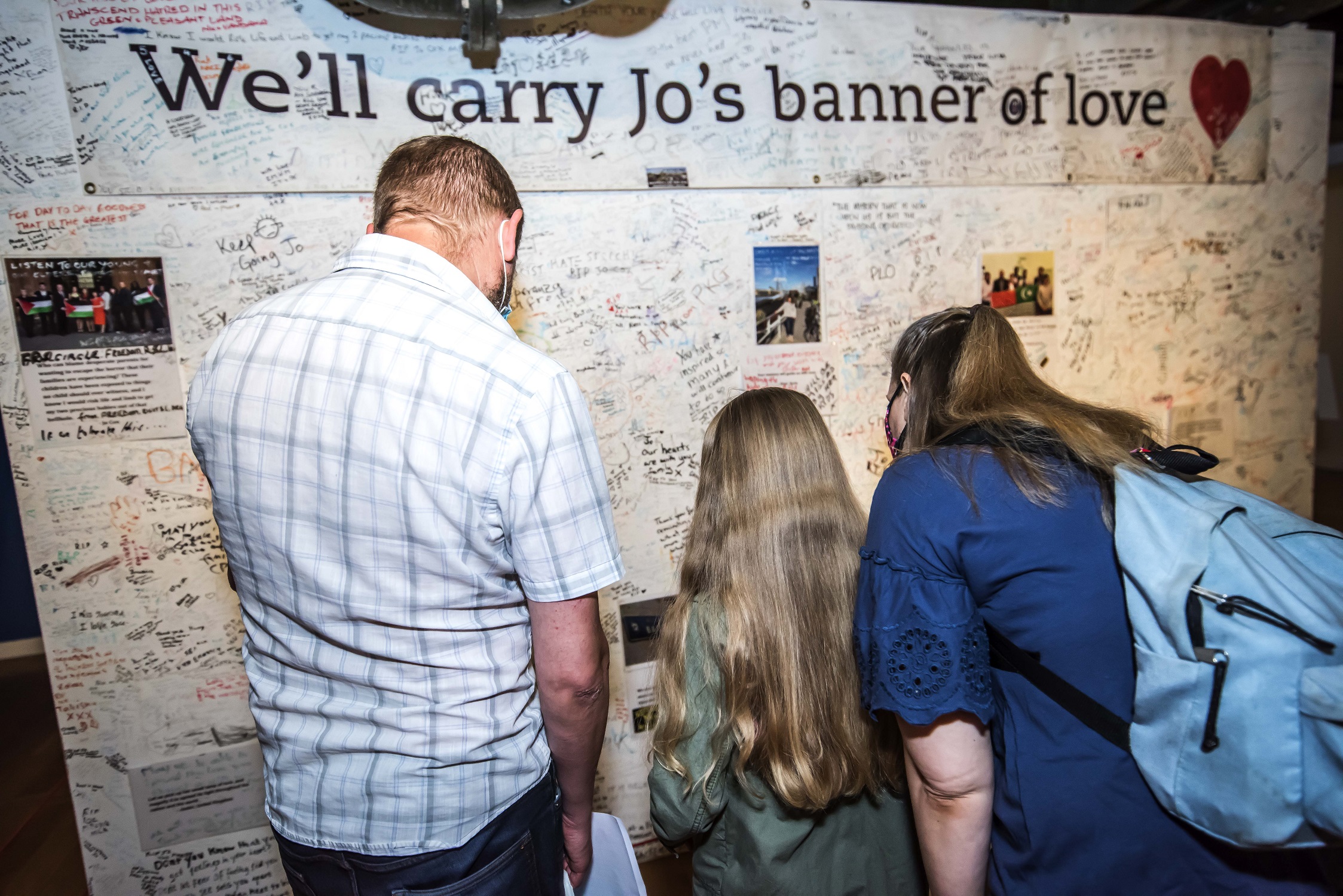 A man, woman and child with long hair face a wall of messages reading 'we'll carry Jo's banner'.