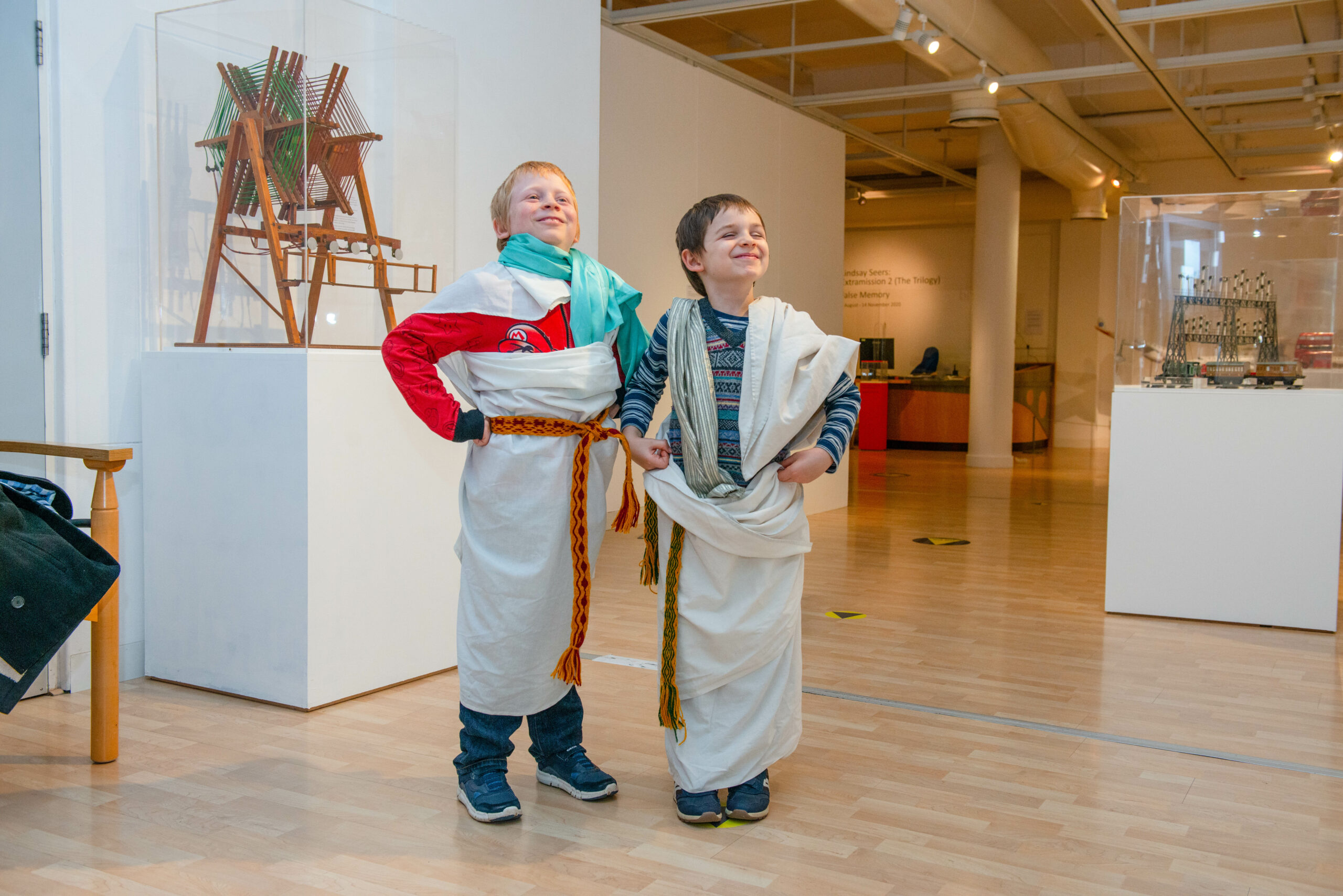 Two children stand posing and smiling in a museum space. They have Roman style clothing items on top of their normal clothes.