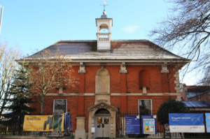 A large red brick museum building with a small tower and weather vane on the roof. There are black railings at the front with banners attached to them.