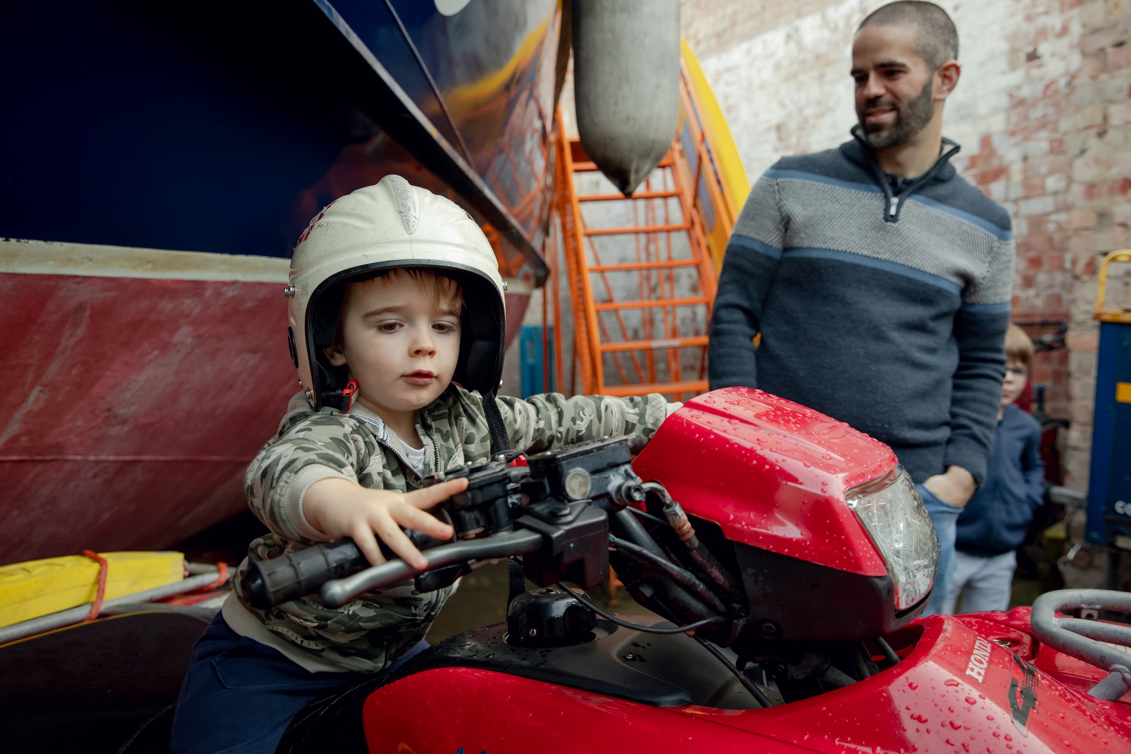 A young boy wears a helmet and sits on a bike at the National Emergency Services Museum. An RNLI boat can be seen in the background. His father grins in the background.