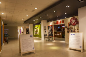 An indoor space with museum interpretation on standing signs and on the walls. On the left is what looks like a shop front with the word 'TAKEAWAY' in big red letters and a Costa Coffee logo.