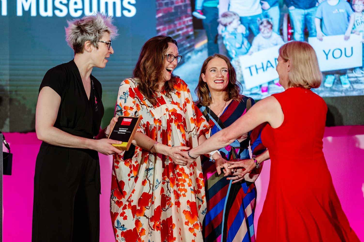 Four women, host Iszi Lawrence and three Kids in Museums trustees, all smiling and wearing dresses stand on stage at the M+H Awards ceremony. Two women hold hands and look delighted, while Iszi holds the Sector Support Award.