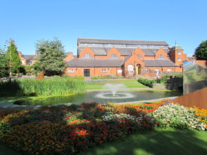 A red brick building sits behind a pond with a fountain in the middle. There are flower beds with red, orange and white flowers.
