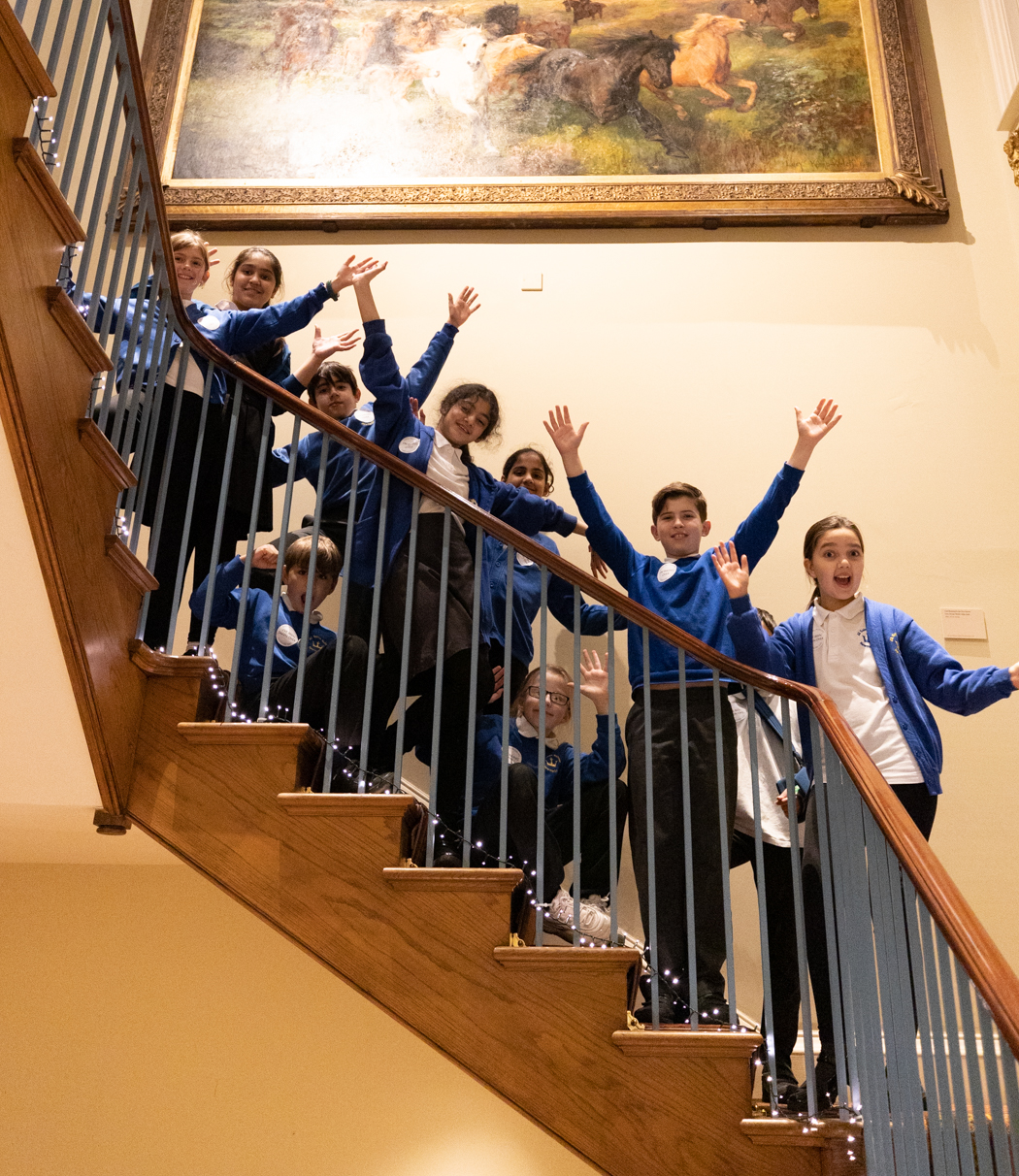 A group of primary school children stand on the stairs at the National Horseracing Museum grinning with their hands in the air.