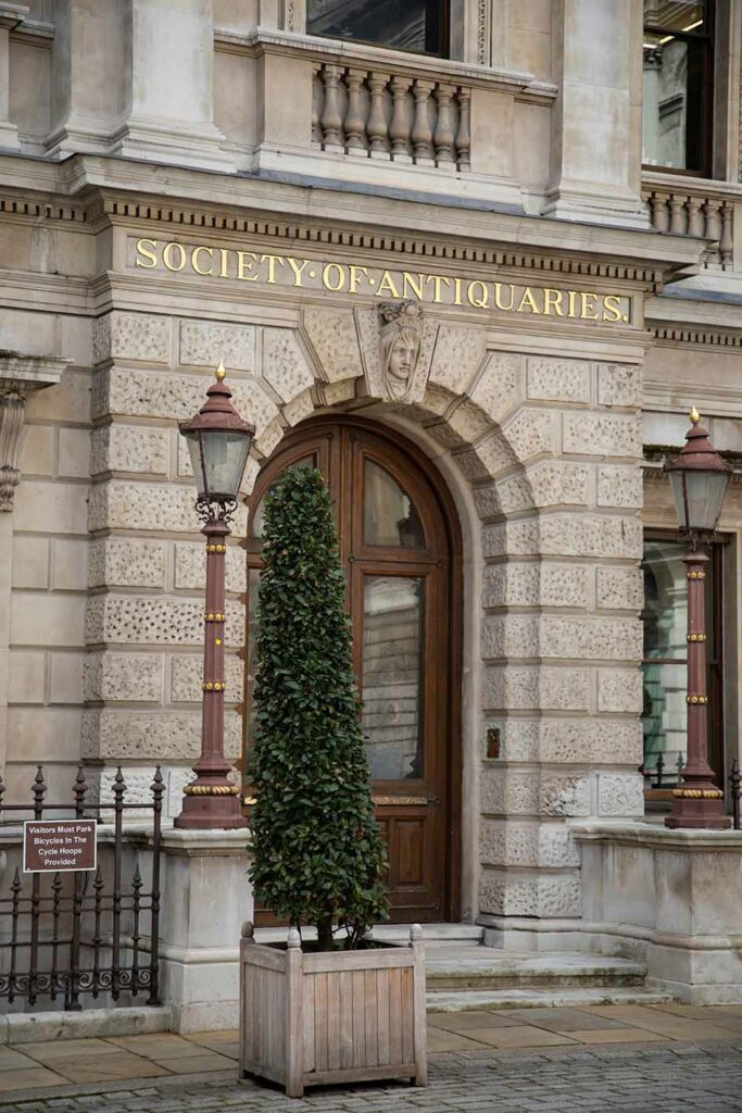 Exterior of Society Of Antiquaries of London. A grand light grey building, featuring a brown door in an arch. Above the door Society Of Antiquaries is written in gold. 