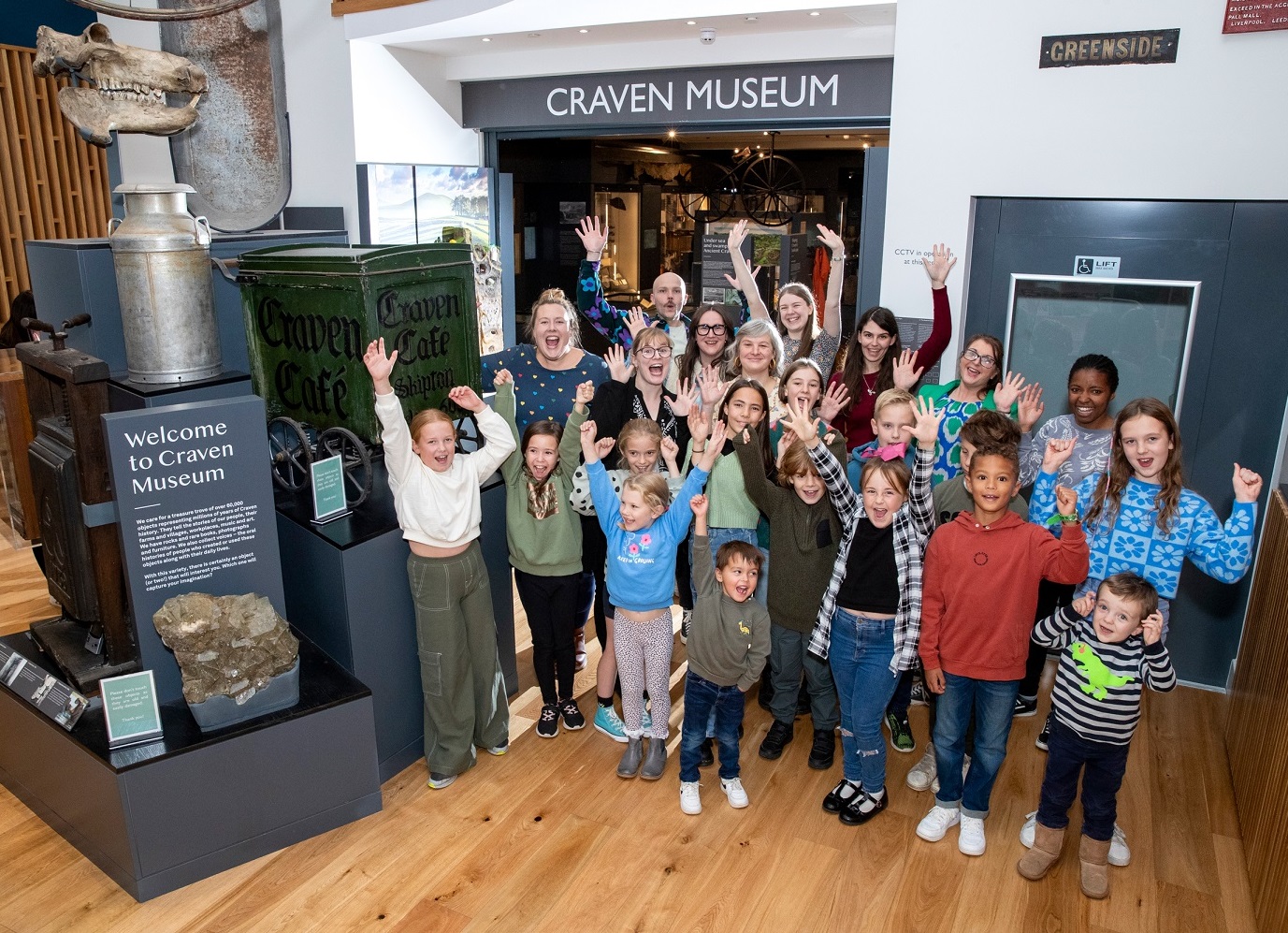 A group of Craven Museum staff and children smile and hold their hands in the air. A sign on the doorway behind them reads Craven Museum and museum displays surround them.