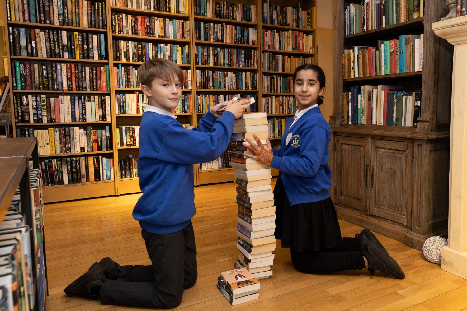 Two primary school children kneel and balance a tall pile of books in the National Horseracing Museum bookshop.