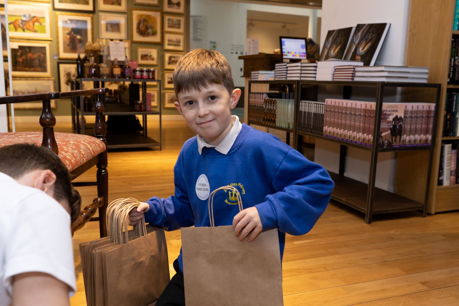 A young boy wearing school uniform and a Takeover Day sticker sorts out paper bags in the sop at the National Horseracing Museum.